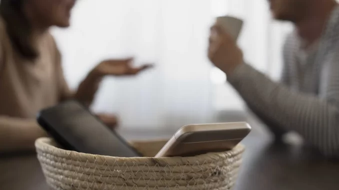 Imagem representando o minimalismo digital com duas pessoas discutindo ao redor de uma mesa e um celular em uma cesta, ilustrando os passos para reduzir a dependência do celular.