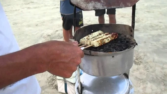 Homem vendendo churrasco e queijo coalho na faixa de areia em Praia Grande, SP, com restrição na venda desses itens na praia. Regras para venda de alimentos na orla.