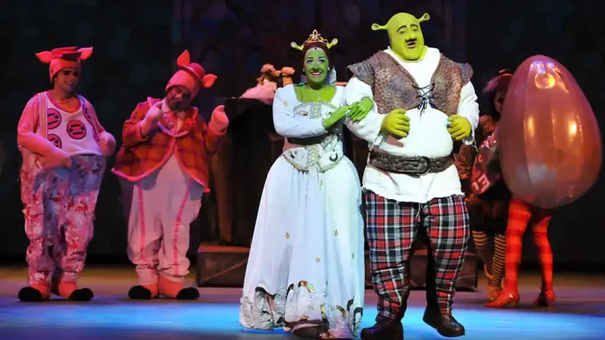Cena do musical Shrek no Teatro Renault com atores em fantasias de personagens do espetáculo, incluindo Shrek e Fiona, com destaque para o trio de estrelas.