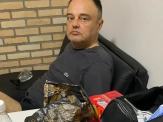 Homem sentado em uma cadeira, ao lado de uma mesa com várias embalagens e objetos, em uma sala com parede de tijolos. Foto relacionada ao STF e acusados de tentativa de explosão no Aeroporto de Brasília.