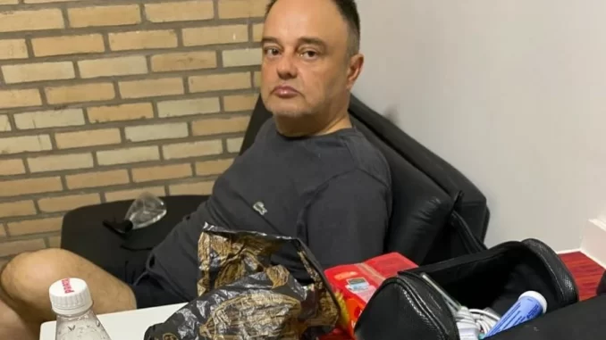 Homem sentado em uma cadeira, ao lado de uma mesa com várias embalagens e objetos, em uma sala com parede de tijolos. Foto relacionada ao STF e acusados de tentativa de explosão no Aeroporto de Brasília.