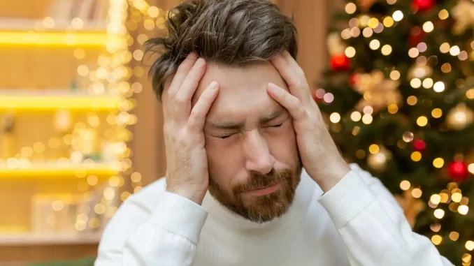 stress-dezembro-cabeca-maos Homem com expressão de estresse e cansaço, segurando a cabeça com as mãos, em ambiente com decoração de Natal, refletindo o estresse de dezembro.