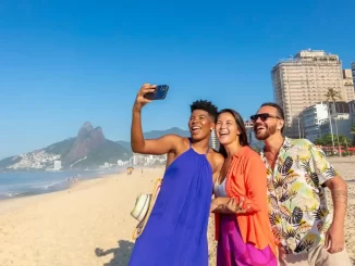 Turistas felizes na praia do Rio de Janeiro celebrando o recorde de visitantes estrangeiros, com vista para o Pão de Açúcar ao fundo e o céu claro.