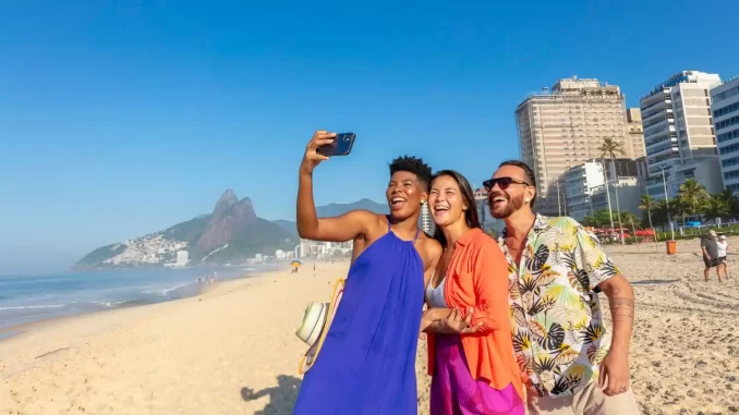 Turistas felizes na praia do Rio de Janeiro celebrando o recorde de visitantes estrangeiros, com vista para o Pão de Açúcar ao fundo e o céu claro.