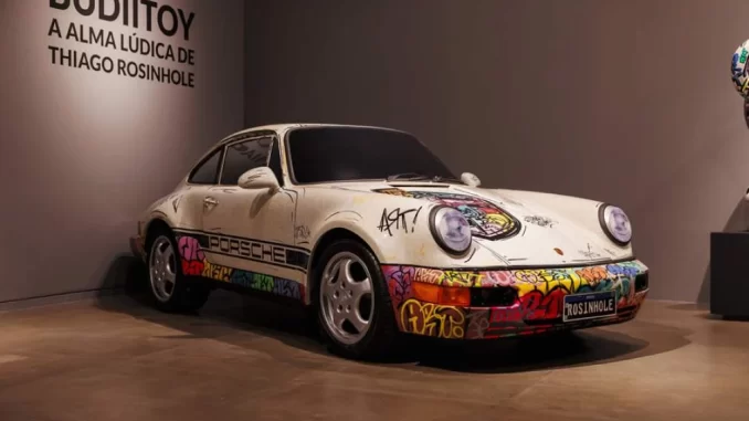 Exposição do carro artística de Thiago Rosinhole na mostra 'Budii: A Alma Lúdica' no Memorial da América Latina em São Paulo, com inscrição 'Budii: A Alma Lúdica de Thiago Rosinhole' na parede.