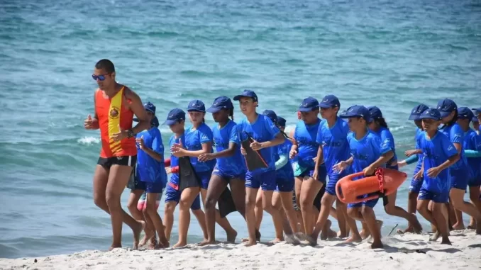 Crianças participando do Projeto Botinho 2026 na praia, vestidas com roupas azuis e usando bonés, com instrutor ao lado, no litoral fluminense do Rio de Janeiro.