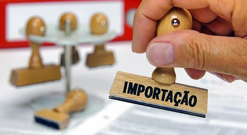 Imagem de uma mão segurando um carimbo com a palavra 'Importação' ao lado de uma balança de teste de produtos e caixas de produtos importados, simbolizando a redução de tarifas de importação pela Camex.