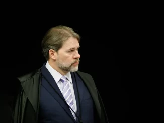 Imagem de um homem com expressão séria, traje formal e cabelo grisalho, aparentando ser uma autoridade judicial, com fundo preto