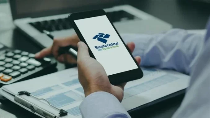 Pessoa verificando o prazo do Imposto de Renda 2026 usando o site da Receita Federal, com documentos organizados ao lado e celular na mão.