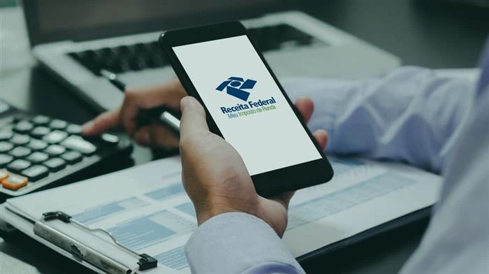 Pessoa verificando o prazo do Imposto de Renda 2026 usando o site da Receita Federal, com documentos organizados ao lado e celular na mão.