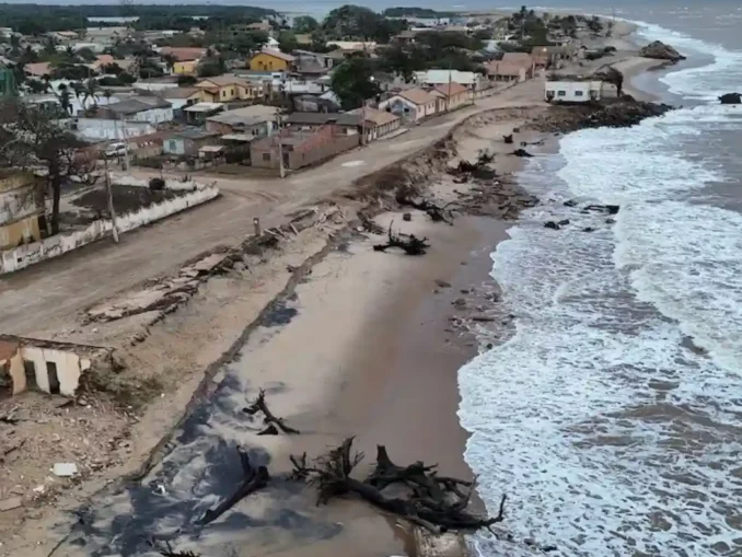 Imagem de uma cidade costeira sendo ameaçada pela elevação do mar, com deslizamentos de terra e árvores destruídas na praia, evidenciando os riscos de desaparecimento até 2030.