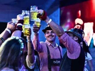 Pessoas brindam com canecas de cerveja durante evento de Oktoberfest, com música ao fundo e clima festivo, em cena no Parque Ibirapuera.