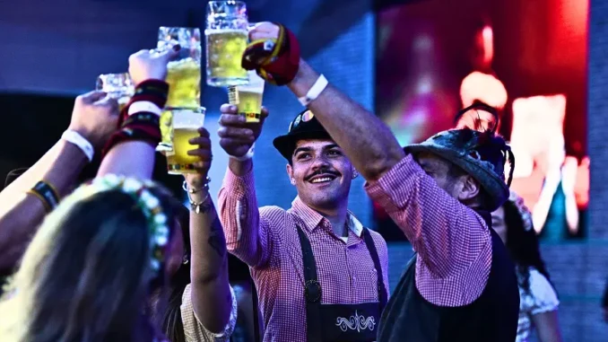 pessoas-brindam-com-canecas-cerveja-oktoberfest-parque-ibirapuera Pessoas brindam com canecas de cerveja durante evento de Oktoberfest, com música ao fundo e clima festivo, em cena no Parque Ibirapuera.