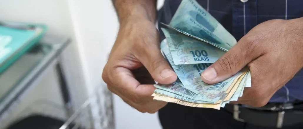 Mãos segurando cédulas de dinheiro, com referência ao salário mínimo que deverá subir para R$ 1.741 em 2027.