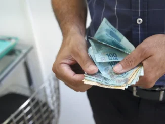 Mãos segurando cédulas de dinheiro, com referência ao salário mínimo que deverá subir para R$ 1.741 em 2027.