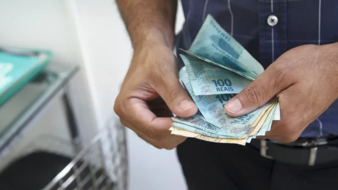salario-minimo-subira-para-1741-em-2027-maos-segurando-dinheiro Mãos segurando cédulas de dinheiro, com referência ao salário mínimo que deverá subir para R$ 1.741 em 2027.
