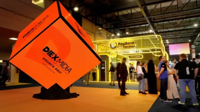 Banner laranja da DIEX MIDIA em feira de negócios no Nordeste, com público circulando em estande ao fundo.