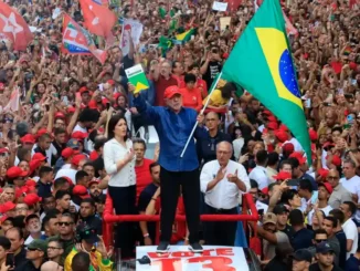 Multidão em manifestação com bandeiras do Brasil, do PT e cartazes, em evento político com Lula em um palanque. Público celebra reeleição presidencial.
