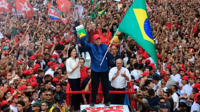 multidao-bandeiras-brasil-pt-lula-reeleicao-presidente Multidão em manifestação com bandeiras do Brasil, do PT e cartazes, em evento político com Lula em um palanque. Público celebra reeleição presidencial.