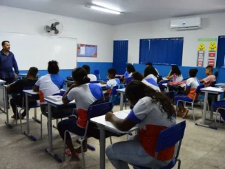 Sala de aula com alunos sentados em carteiras e um professor à frente, em ambiente escolar com ventilação e ar-condicionado, mostrando rede pública de ensino