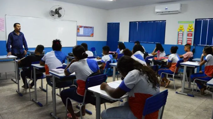 sala-de-aula-alunos-professor-rede-publica-ensino-sp Sala de aula com alunos sentados em carteiras e um professor à frente, em ambiente escolar com ventilação e ar-condicionado, mostrando rede pública de ensino